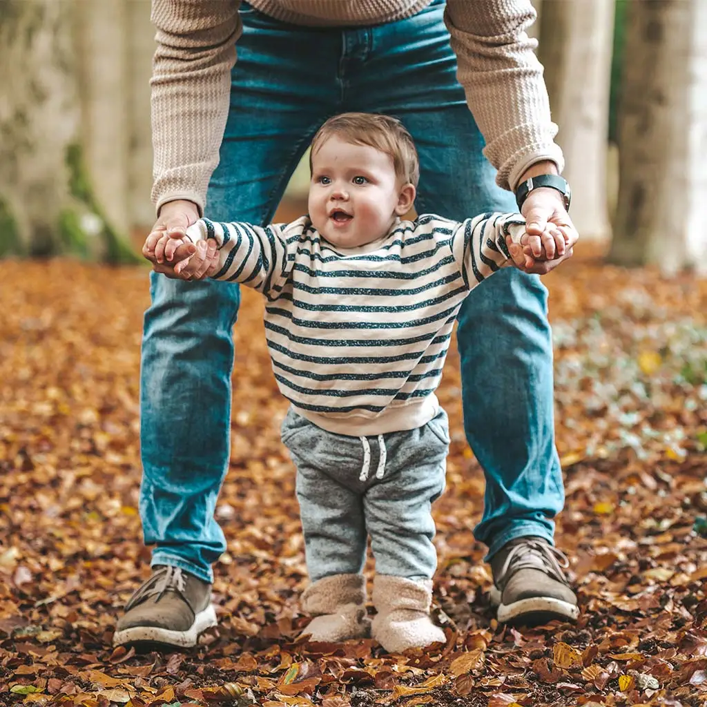 Kind loopt tussen de benen van de zijn vader terwijl ze elkaars handen vasthouden tijdens de familieshoot in het bos.