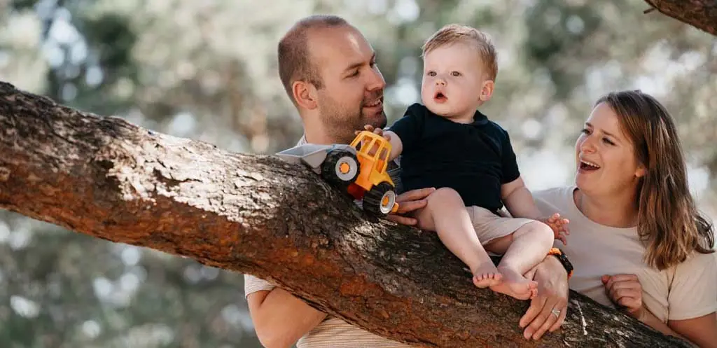Kind zit op een boomstam met een speelgoedauto, ouders staan liefdevol achter het kind tijdens de familieshoot op het Wekeromse Zand